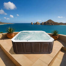 Cargar imagen en el visor de la galería, jacuzzi cuadrado blanco con hidromasaje y faldón imitación madera, en una terraza exterior lujosa con vista a la playa